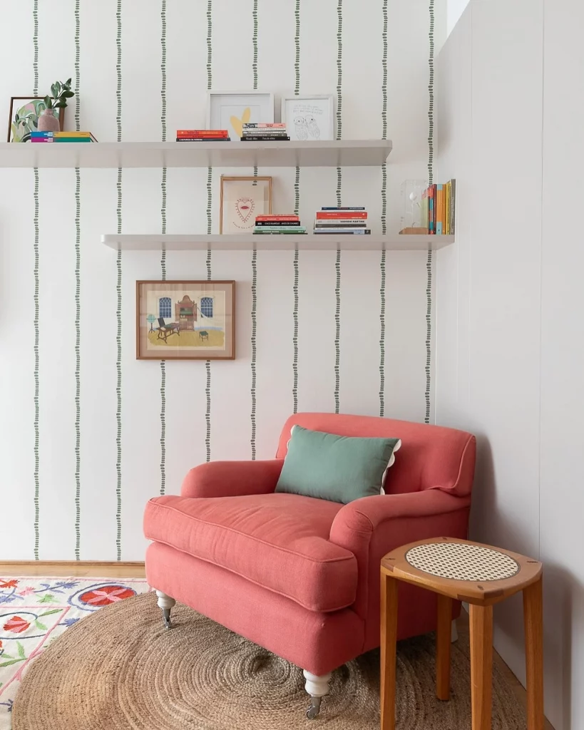 Espaço de leitura com poltrona cor de goiaba confortável, almofada verde, prateleiras com quadros e livros, papel de parede listrado.