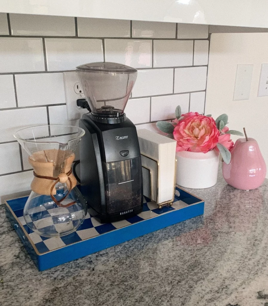 Bandeja xadrez azul com guardanapos, máquina de café e coador de café, tem um vaso de flor pequeno ao lado.