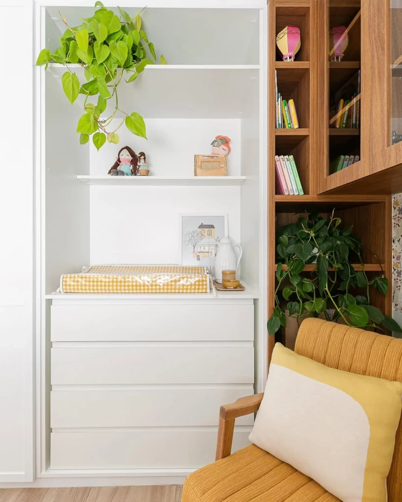 Cantinho de quarto infantil com trocador, poltrona amarela, prateleiras com itens decorativos e planta.