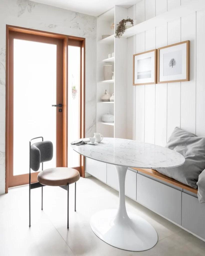 Canto de parede texturizado e branco, com mesa oval branca, prateleiras e quadro.