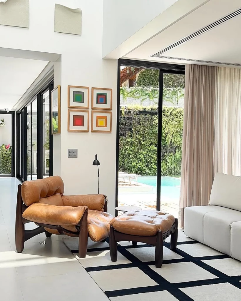 Quadros quadrados com diferentes cores, poltrona de couro marrom, porta de vidro e sofá claro.