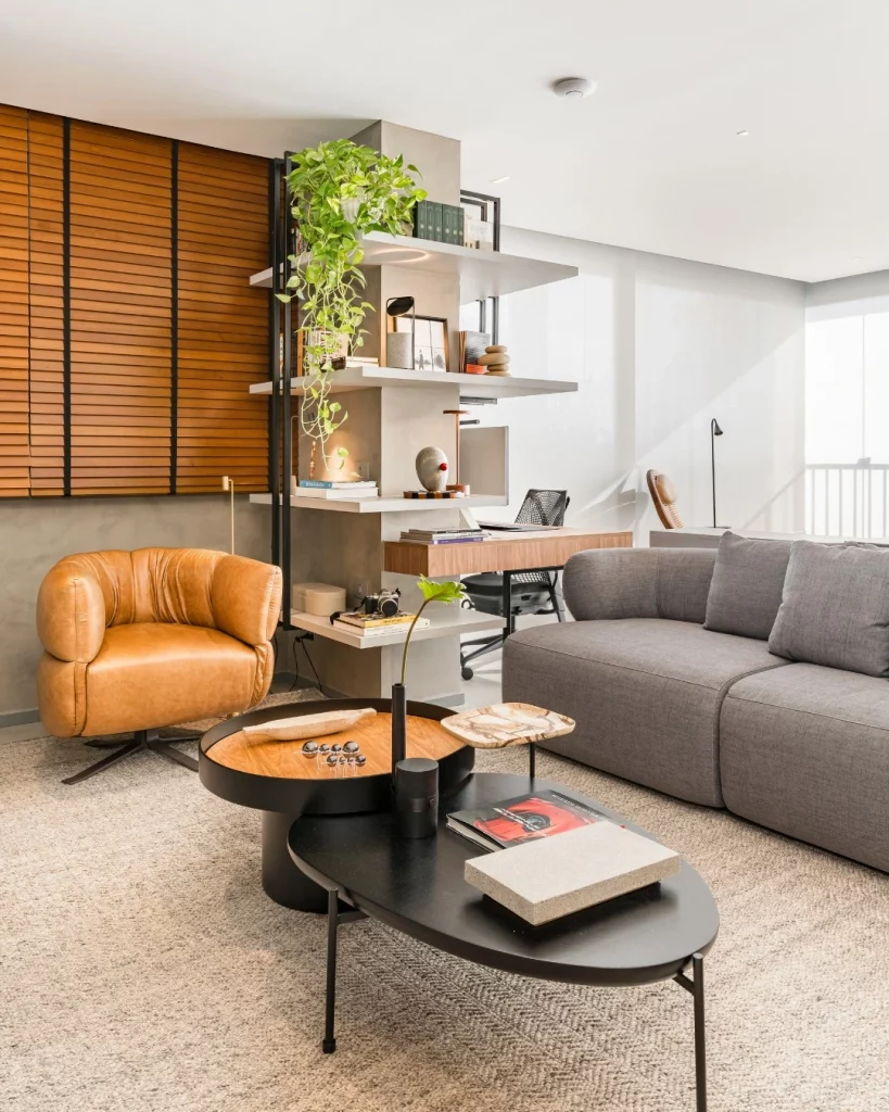 Planta em prateleiras cantoneiras, sofá cinza, poltrona de couro marrom caramelo claro e mesa de centro na sala.