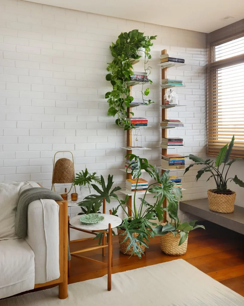 Canto com muitas plantas no chão, estante de livros diferente, sofá e mesa lateral.