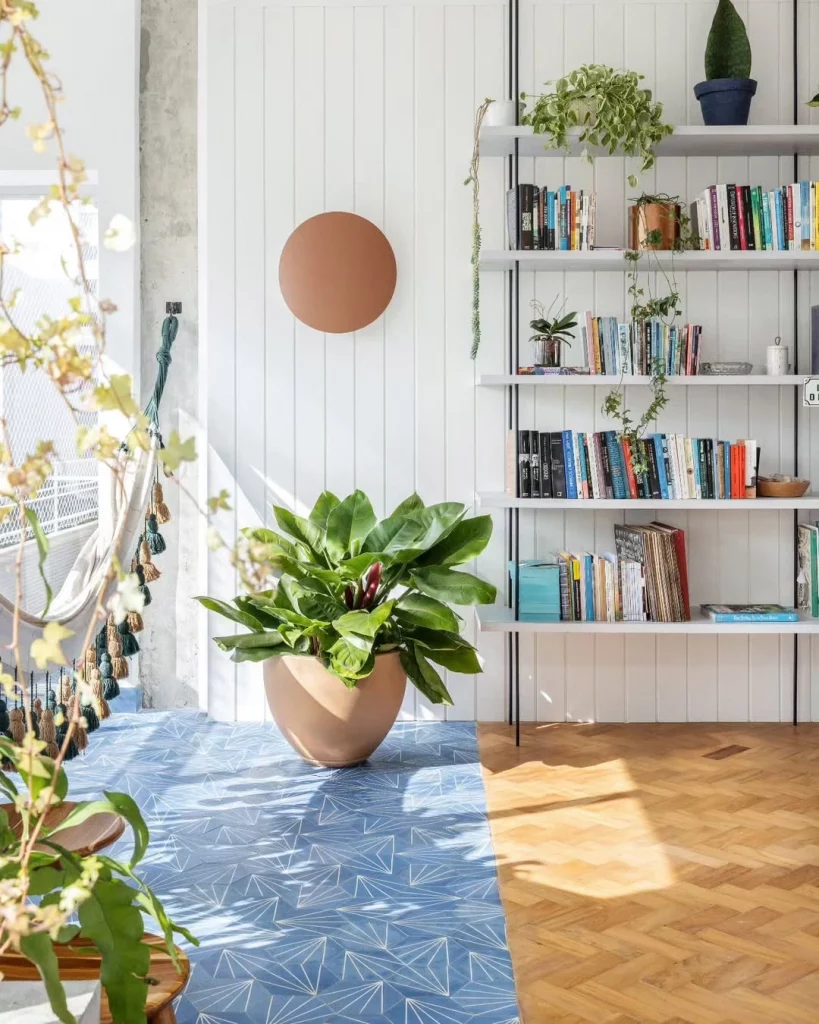 Cantinho da leitura com estante e livros, vaso de plantas, escultura redonda na parede e tapete azul.