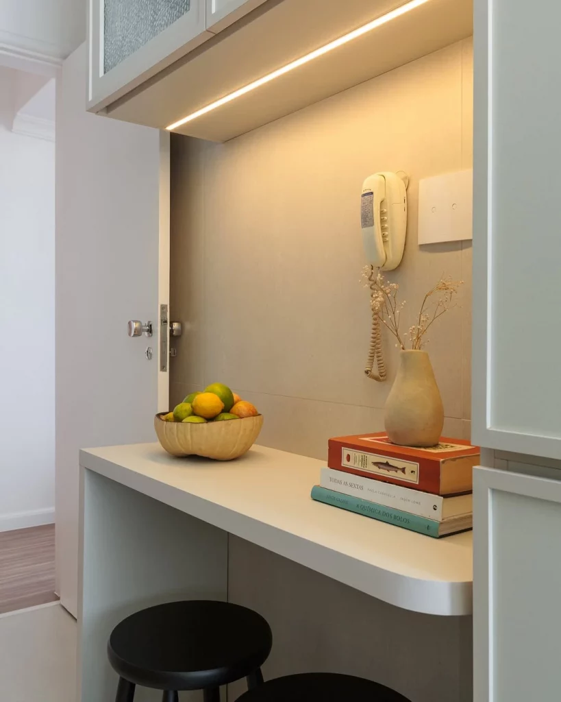 Canto iluminado com luz de LED amarela, interfone, livros decorativos, fruteira e mesa cantoneira.