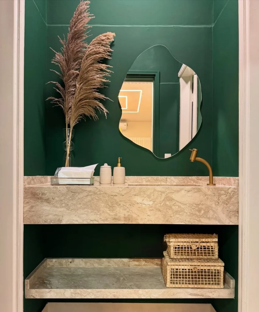 Lavabo com parede verde escuro, flor seca grande na pia, espelho orgânico e caixas de palha.