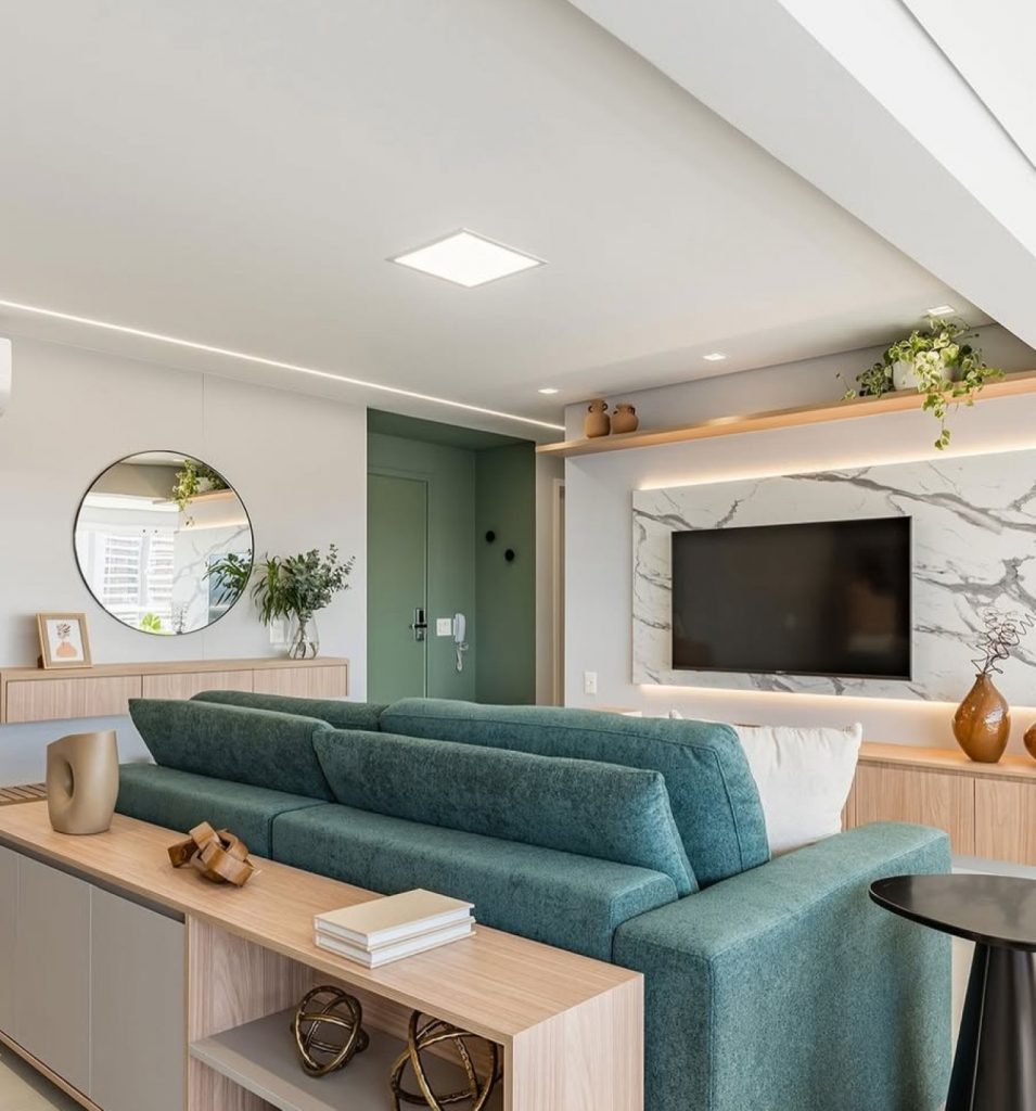 Sala de estar com iluminação geral no teto, sofá verde, televisão, aparador e espelho na parede lateral.