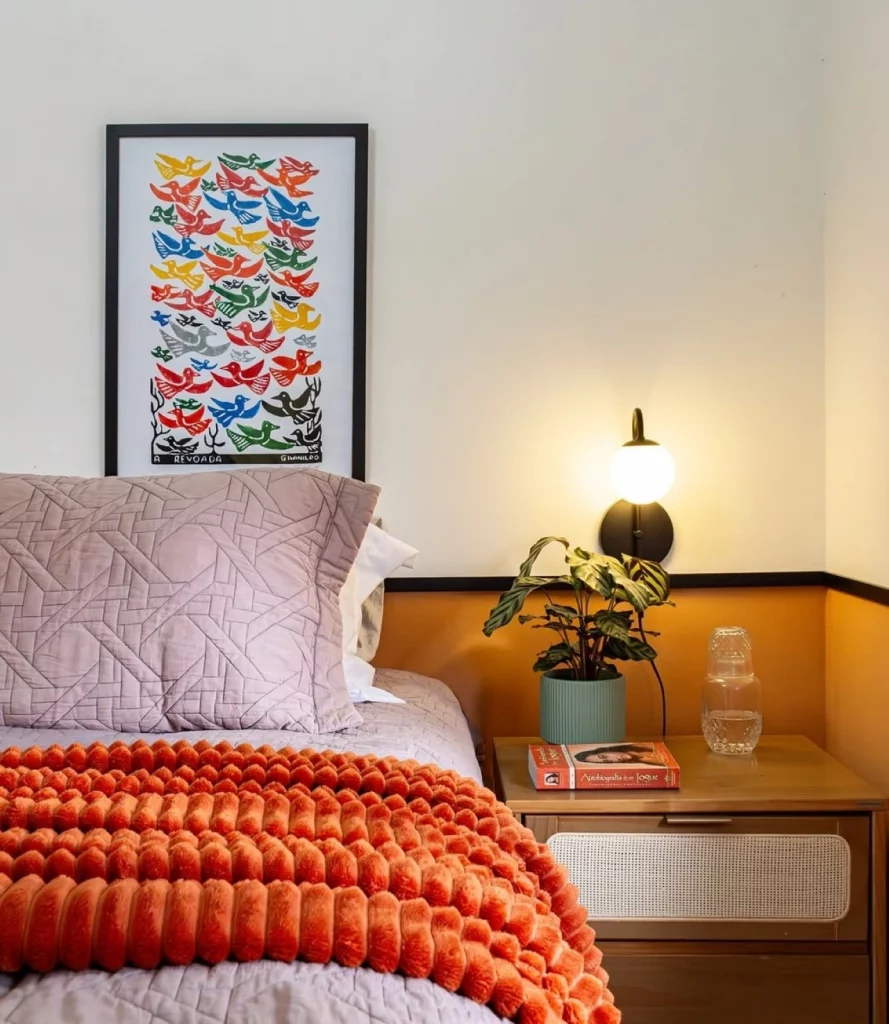 Iluminação quente no quarto em arandela ao lado da cama, mesa de cacbeceira, quadro na parede e manta terracota.
