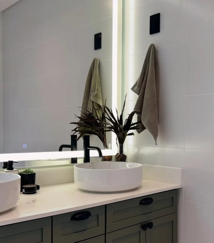 Espelho com iluminação branca de LED, banheiro com toalha cinza pendurada ao lado da pia.