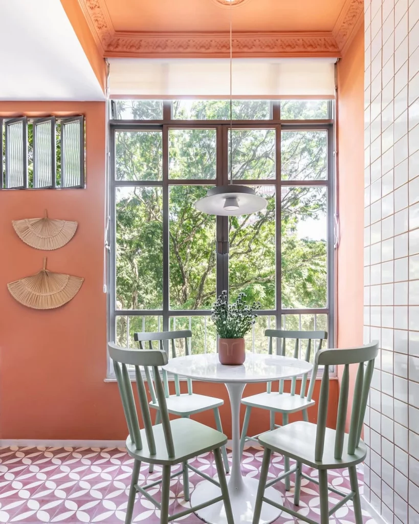Cantinho aconchegante decorado com leques de palha, parede terracota e mesa com cadeiras verdes.