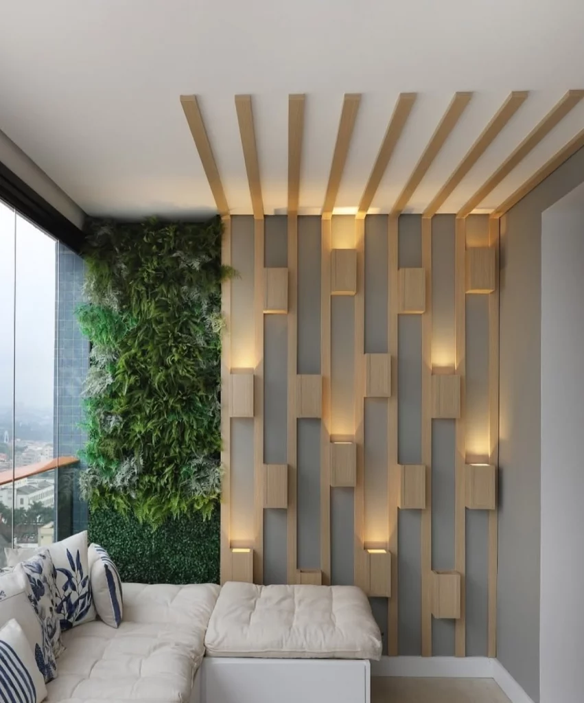 Iluminação decorativa na varanda de apartamento, com arandelas de madeira e luz amarela, parede verde de plantas ao lado.