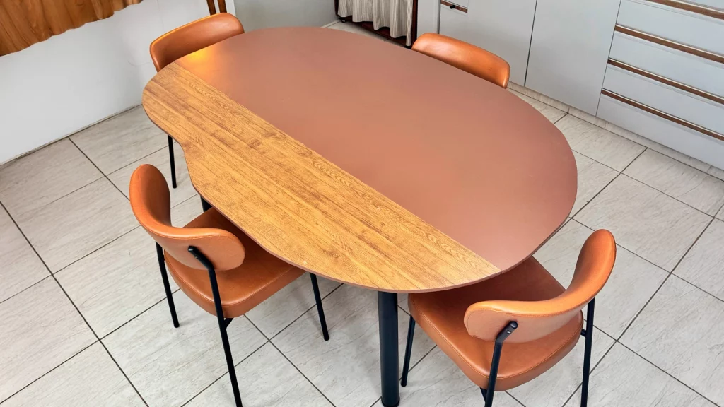 Mesa de jantar de madeira, com tampo orgânico pintado de marrom apenas pela metade e cadeiras com assento de couro.