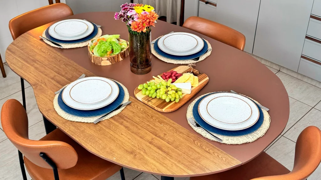 Mesa de jantar em formato orgânico com jogo americano, pratos, vaso de flores e tábuas de frios.