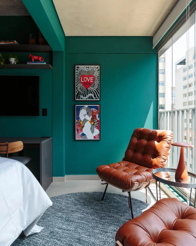 Parede verde com dois quadros e uma poltrona marrom de couro próxima.