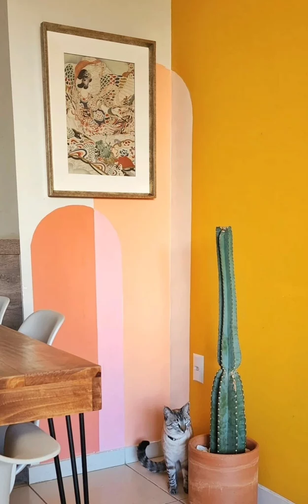 Pintura no canto da parede, com tons quentes de rosa, amarelo e laranja, cacto e quadro compondo o restante da decoração.