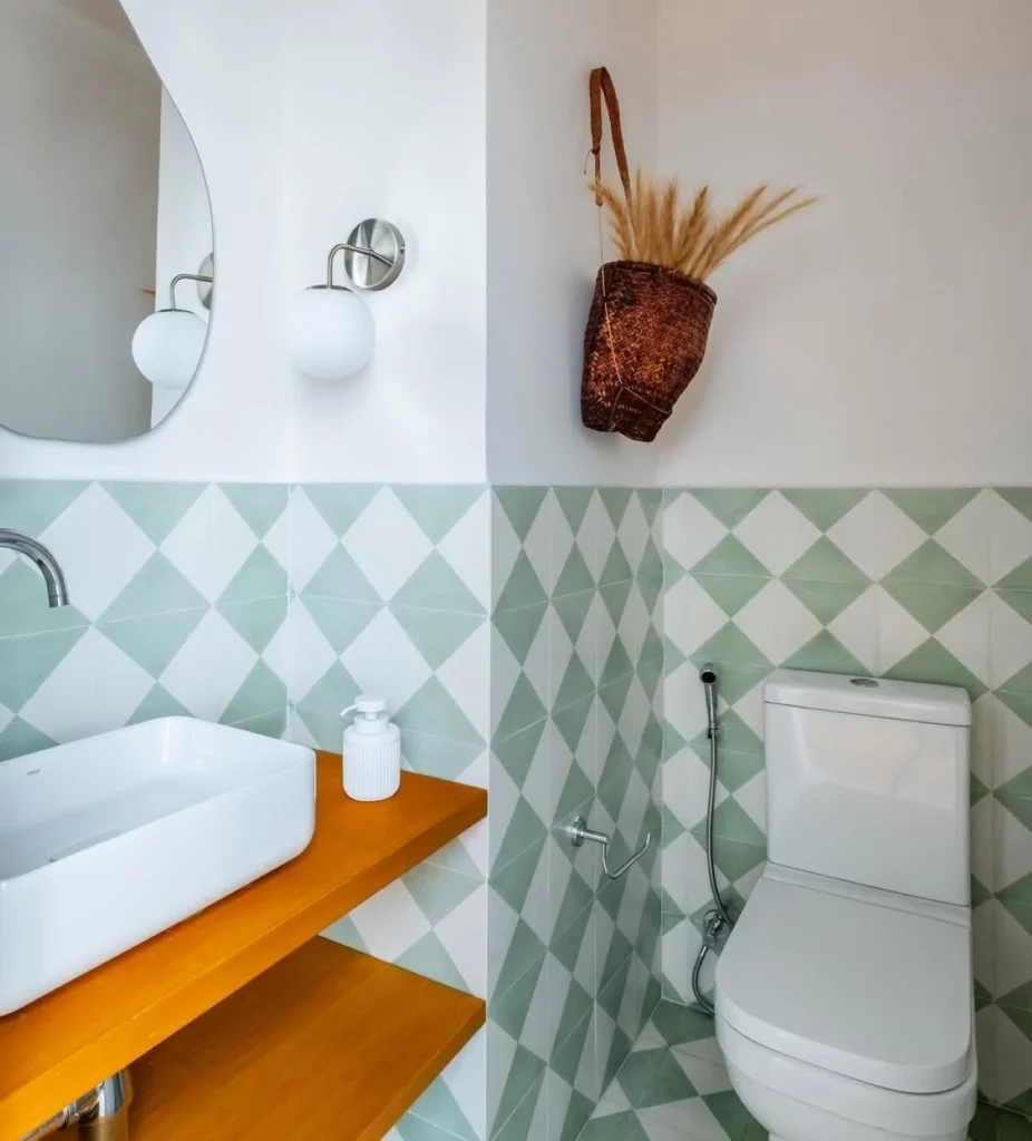 Banheiro com vaso de ramos secos pendurado na parede, cerâmica xadrez de verde e branco, madeira envernizada na pia.