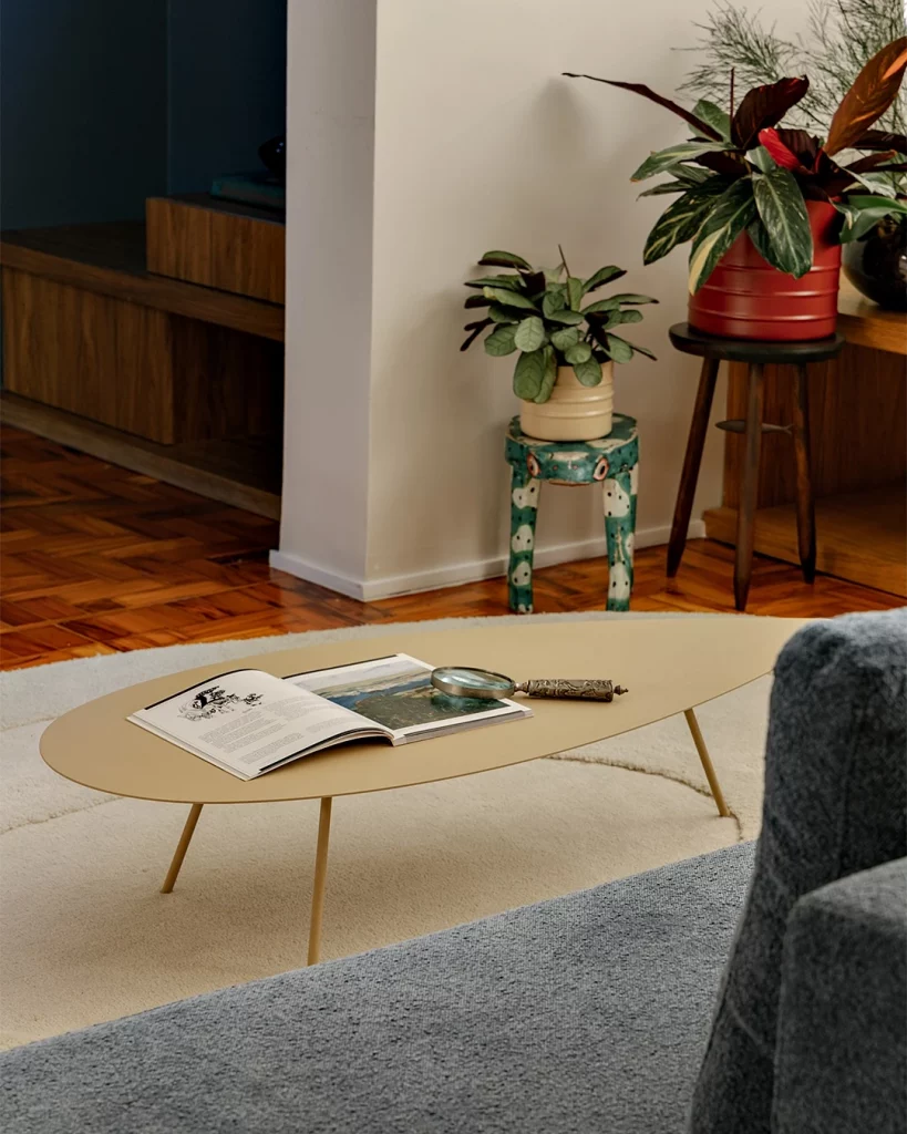 Mesa de centro bege amarelado em formato orgânico, com bancos e plantas no fundo.