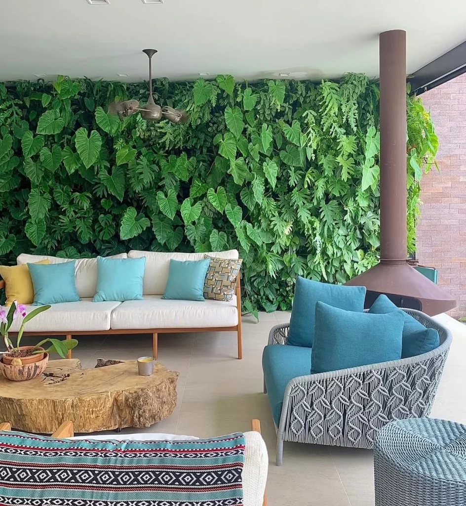 Área externa de casa com sofás e almofadas azuis, parede verde de folhas no fundo.