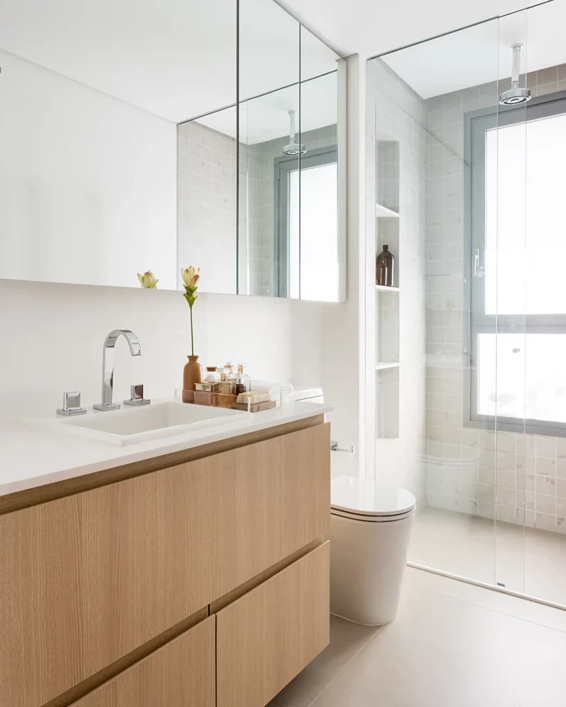 Banheiro clean e suave, com armário de madeira e porcelanato acetinado branco, espelho grande acima da pia.
