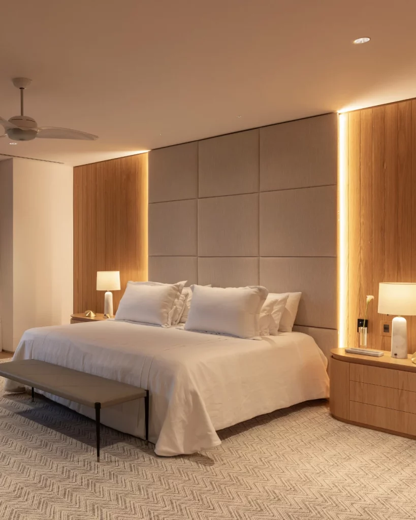 Cabeceira de cama que vai até o teto, modular e estofada em tom neutro, com iluminação nas laterais.