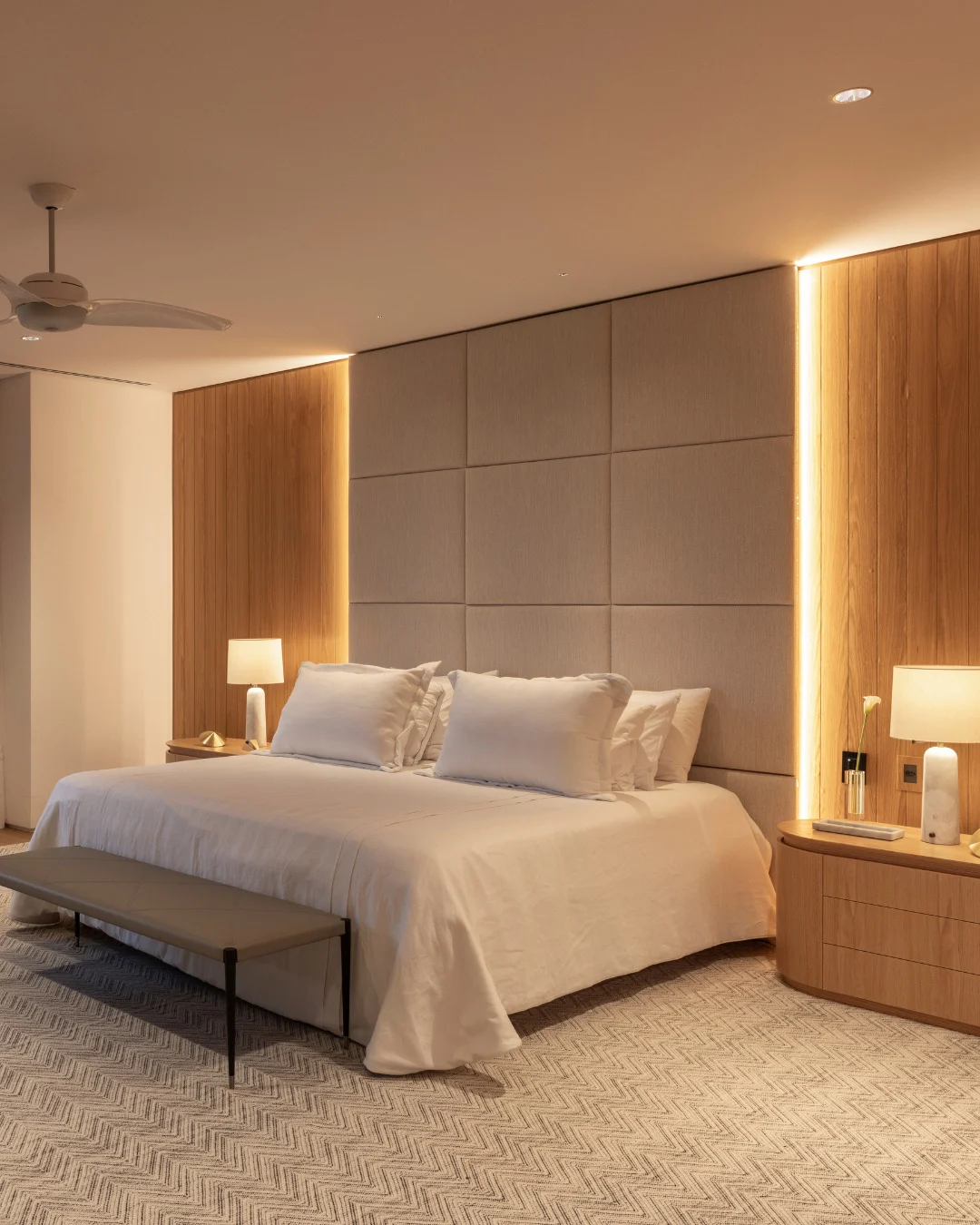 Cabeceira de cama que vai até o teto, modular e estofada em tom neutro, com iluminação nas laterais.