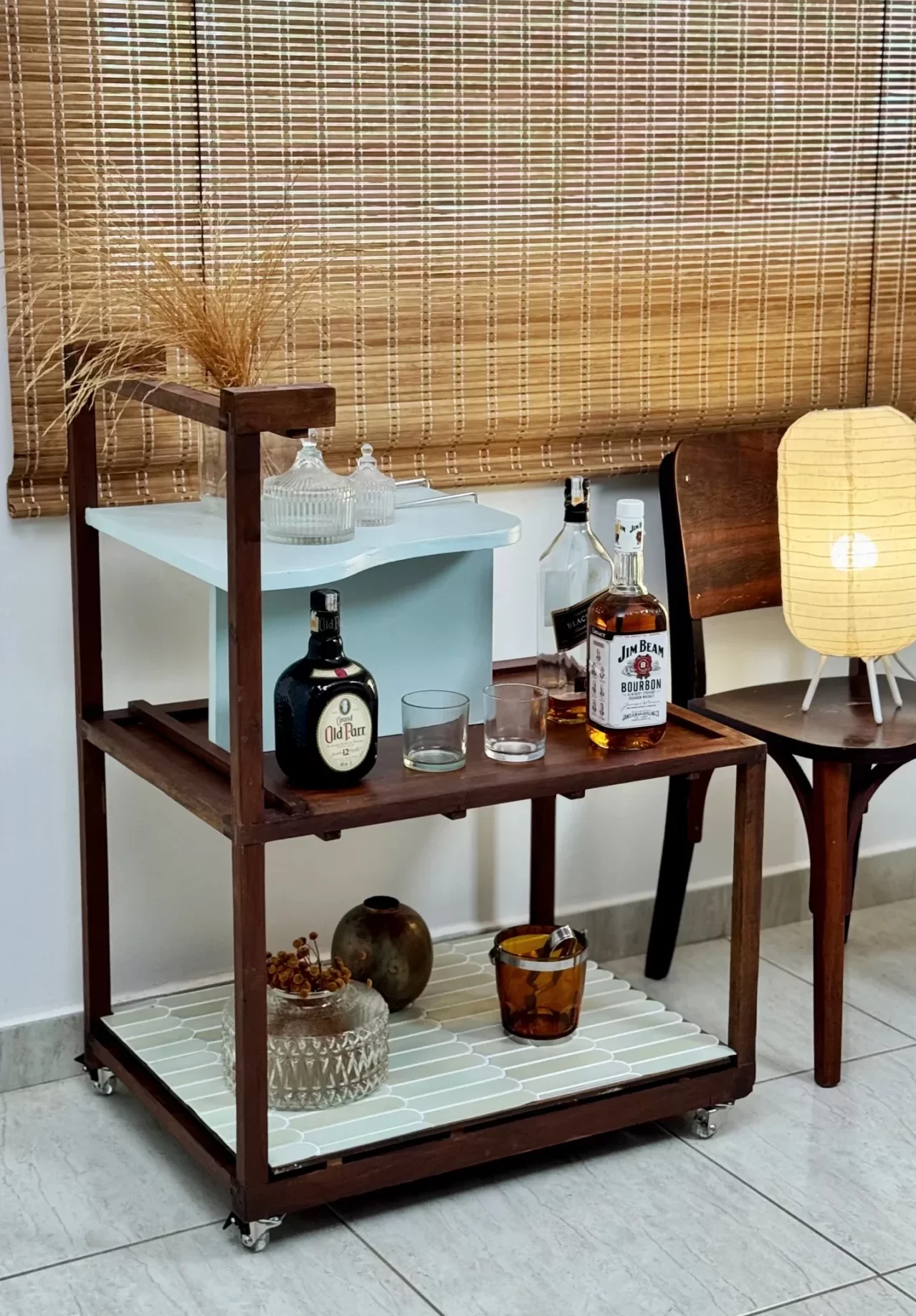 Carrinho-bar com bebeidas, feito de madeira, com luminária na mesa lateral ao lado.