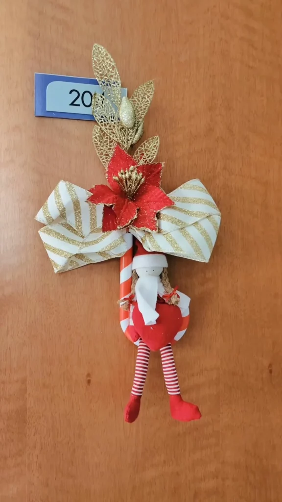 Enfeite de porta natalino com boneca de pano, doce listrado, flor de natal e laço listrado bege com branco.