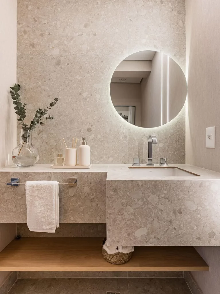 Prateleira de banheiro de madeira, vaso de vidro na pia e espelho iluminado.