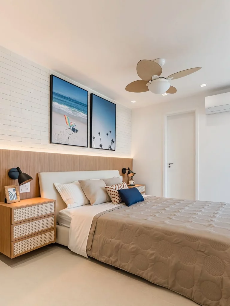 Quarto com quadros de paisagem azuis na parede, cabeceira iluminada, mesa lateral e roupa de cama bege.