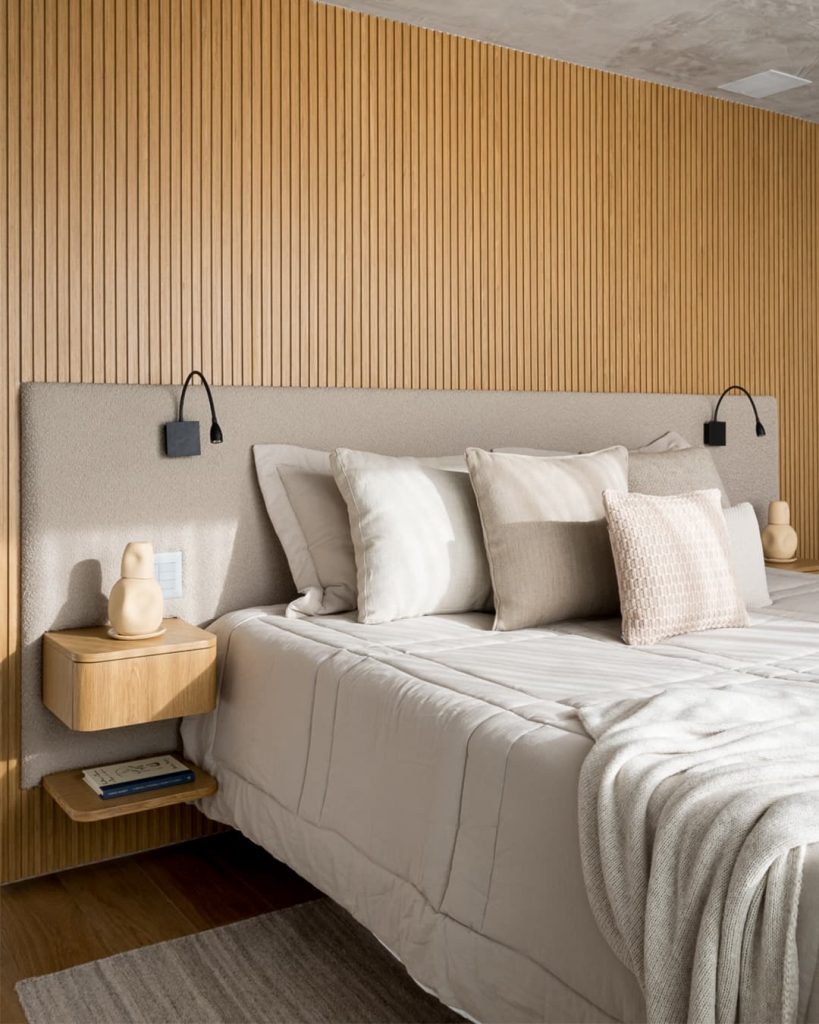 Quarto com cabeceira cinza, arandelas e mesa lateral texturizada.