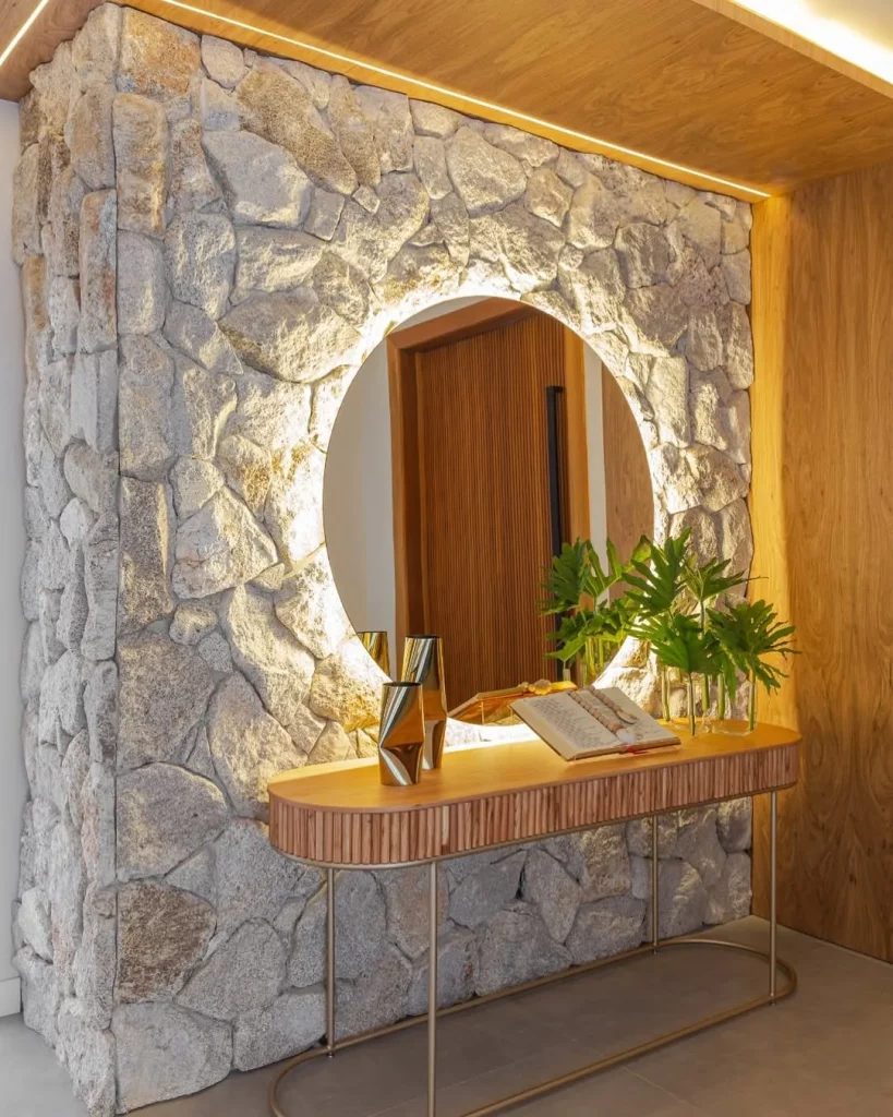 Hall de entrada com espelho iluminado com fita de led, parede de pedra e aparador.