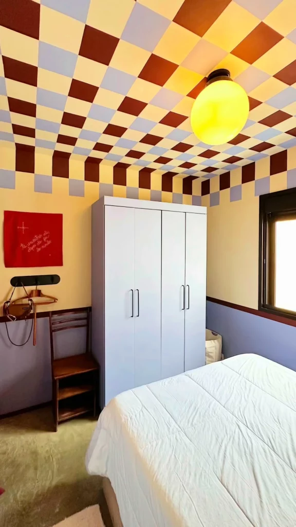 Quarto com quadro vermelho, teto pintado em xadrez e guarda-roupas com pintura branca e puxadores pretos.