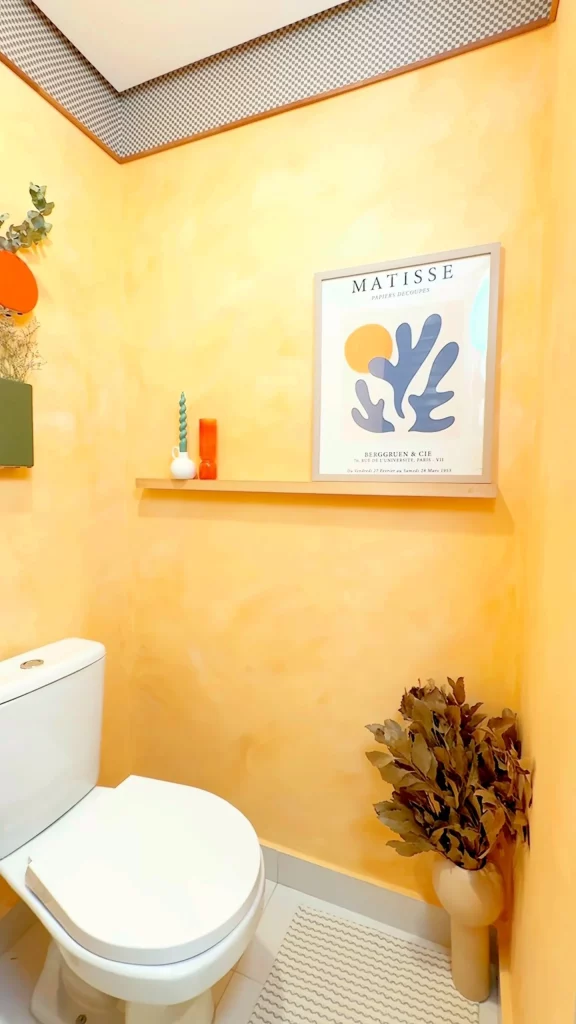 Parede de lavabo com prateleira, pintura amarela, quadro e planta no chão.