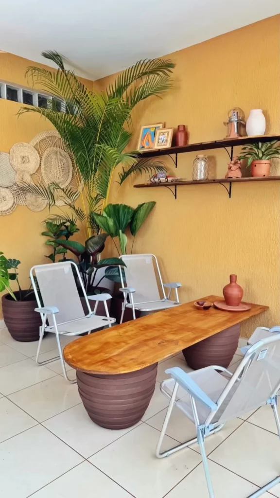 Varanda de tema brasilidade, com mesa no centro feita com madeira e vasos de planta, cadeiras de praia, prateleiras e parede amarela.