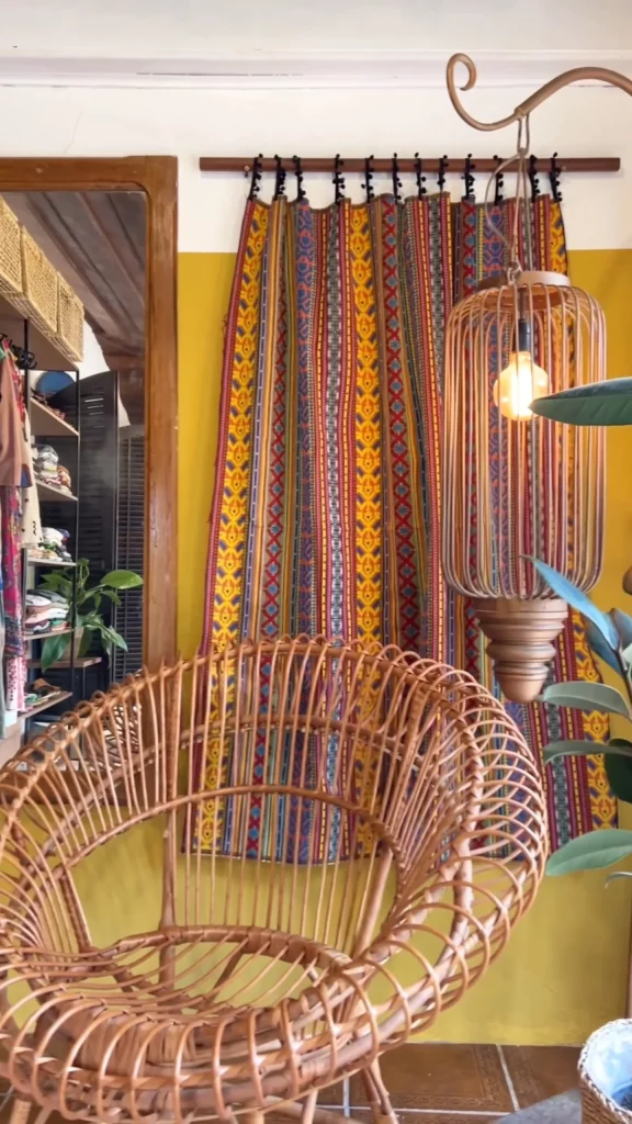 Parede amarela com tecido pendurado, poltrona de fibra natural e luminária.