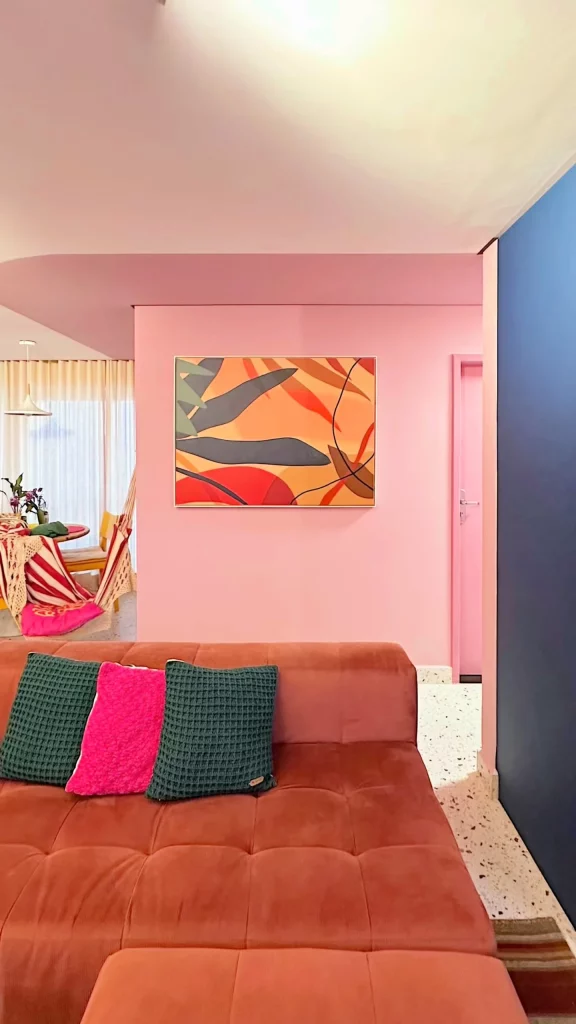 Parede pintada de rosa com quadro grande na parede, sofá amarronzado e almofadas coloridas.