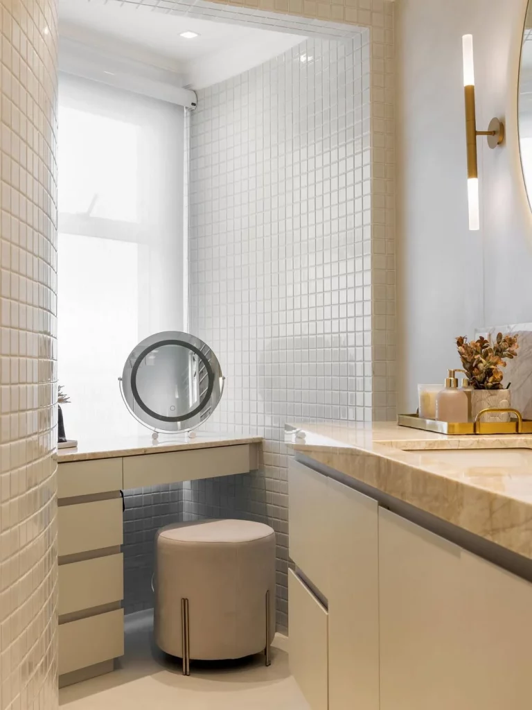 Penteadeira no banheiro de frente para a janela para aproveitar a luz natural, puff neutro e armário com detalhes marrons.