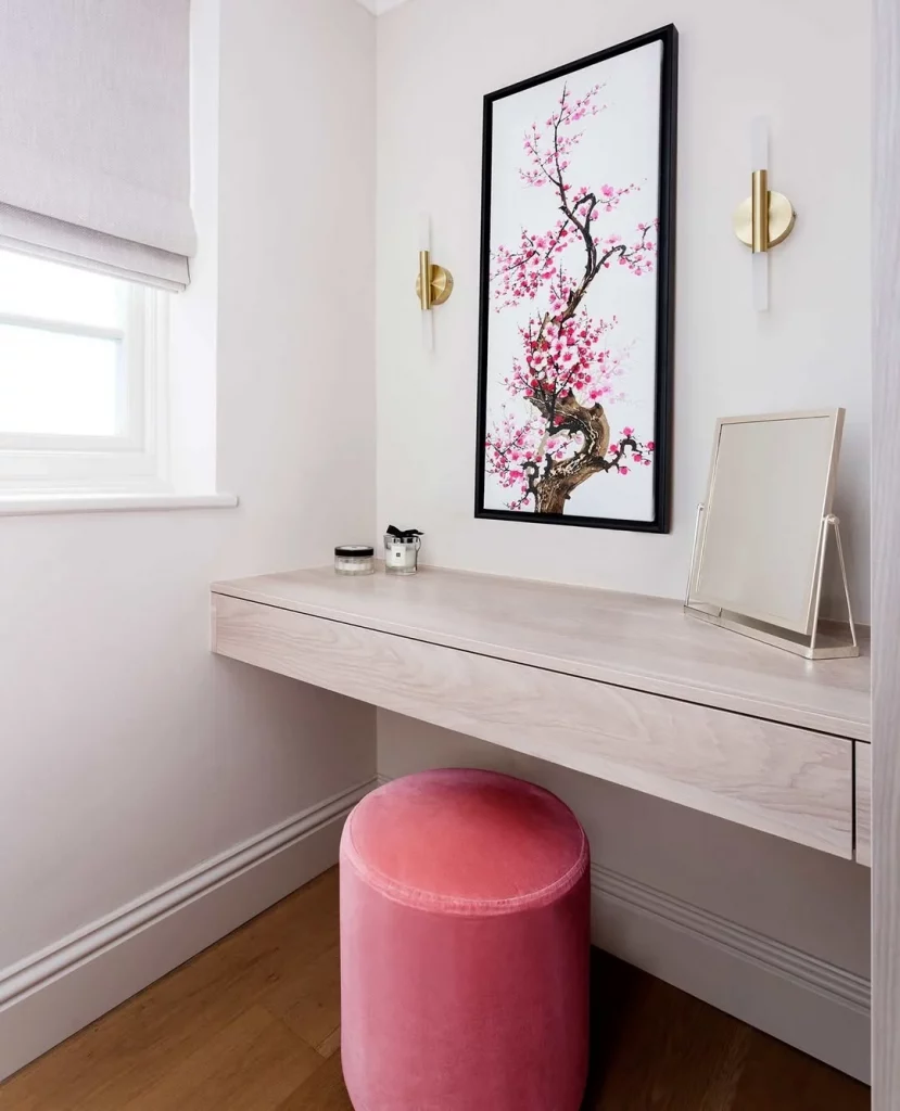 Penteadeira no cantinho perto da janela com quadro, espelho na mesa e puff rosa.