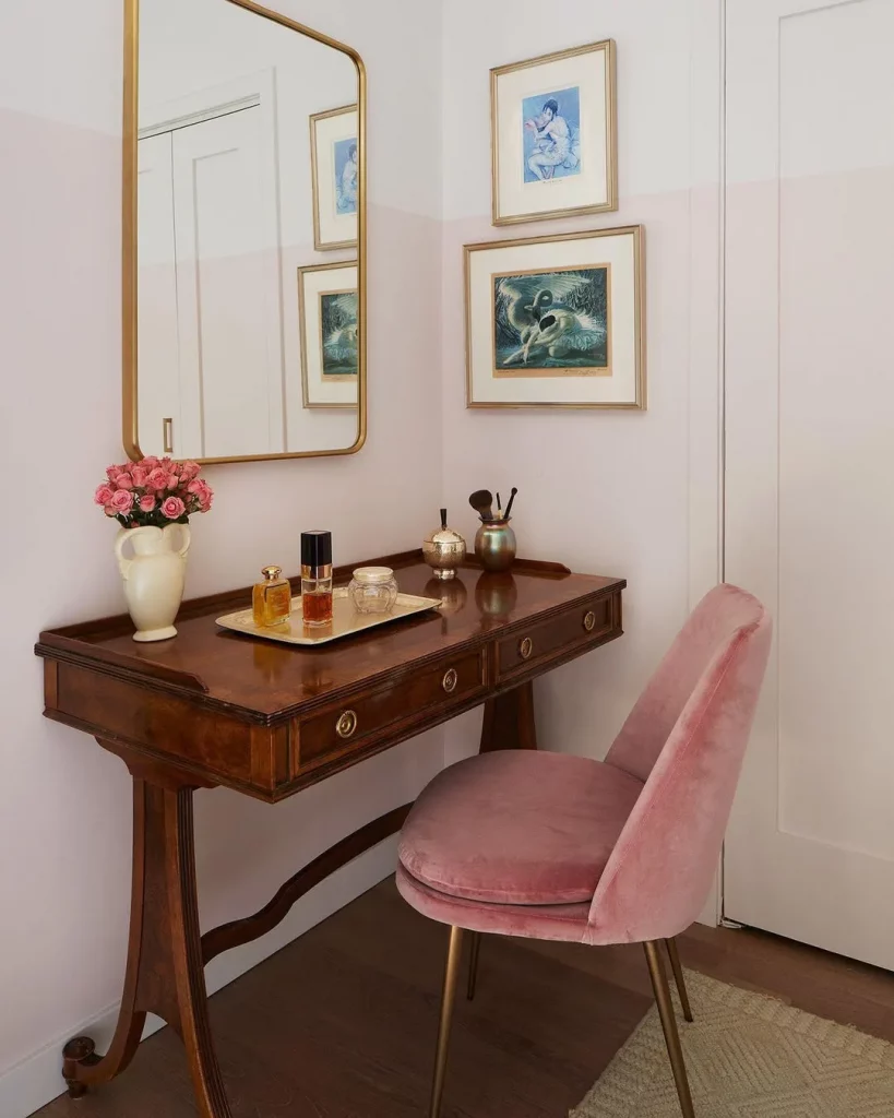 Penteadeira com toque de modernidade e retrô, feita de madeira, com cadeira rosa de veludo e quadros ao lado.
