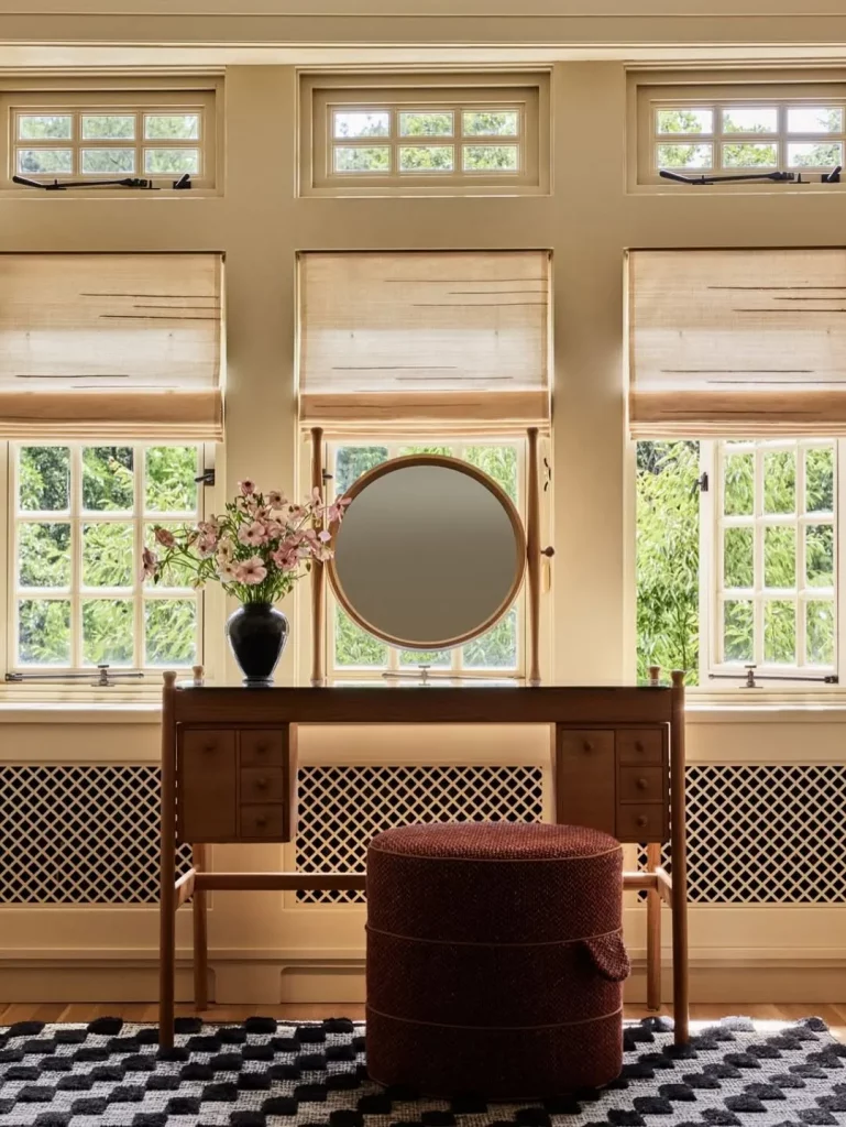 Penteadeira retrô com espelho redondo, gavetas e de frente para muitas janelas do quarto.