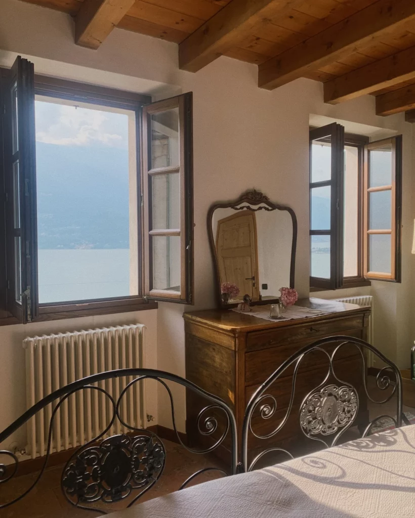 Penteadeira com espelho bem trabalhado, de madeira, perto das janelas e da cama.