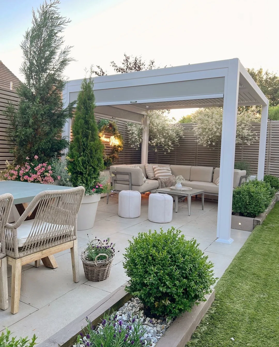 Pergolado branco na área externa elegante, com plantas, mesa e sofás.