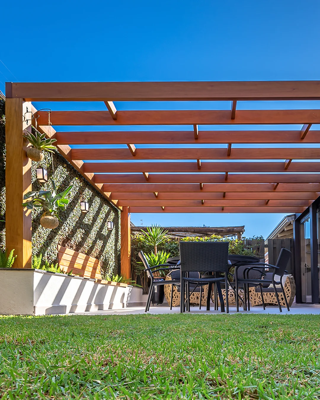Pergolado de madeira, grama e cadeiras pretas.