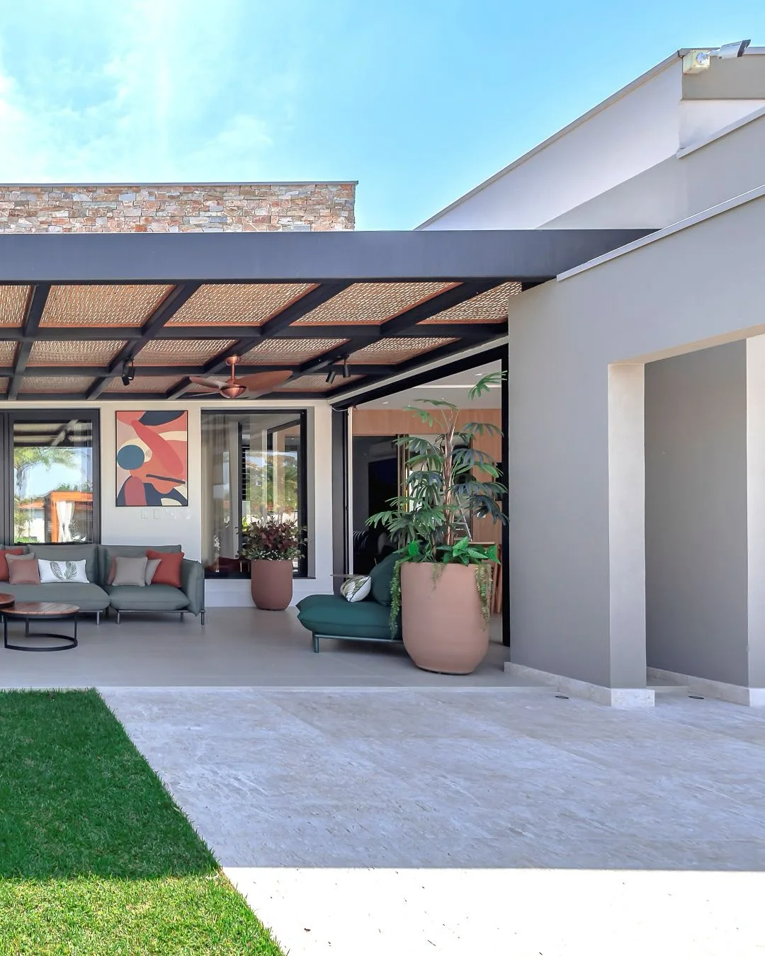 Pergolado preto na varanda, gramado e piso cinza, vaso de planta, quadro na parede e sofá.