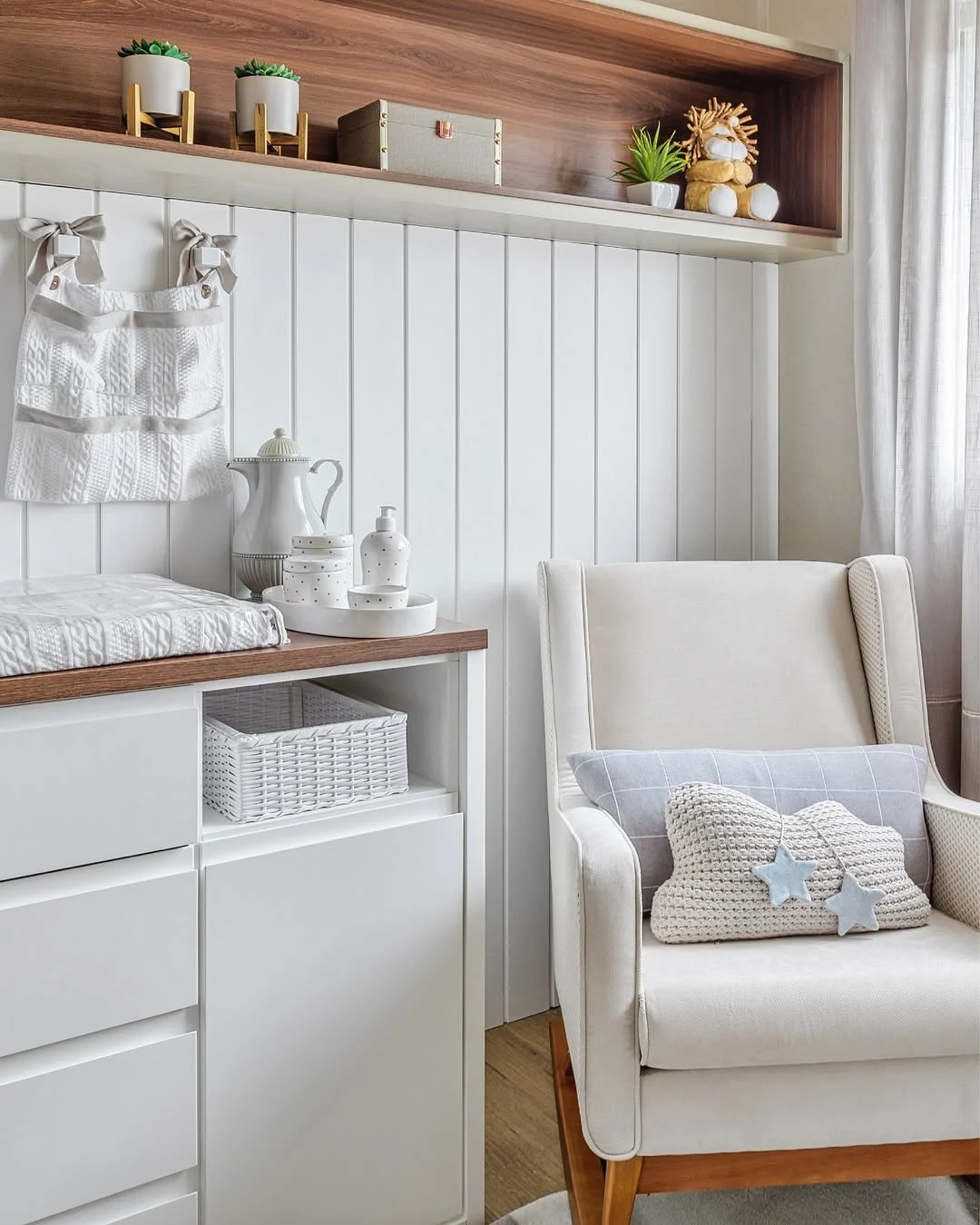 Cantinho do quarto de bebê com poltorna de amamentação, almofada de nuvem e cômoda branca.