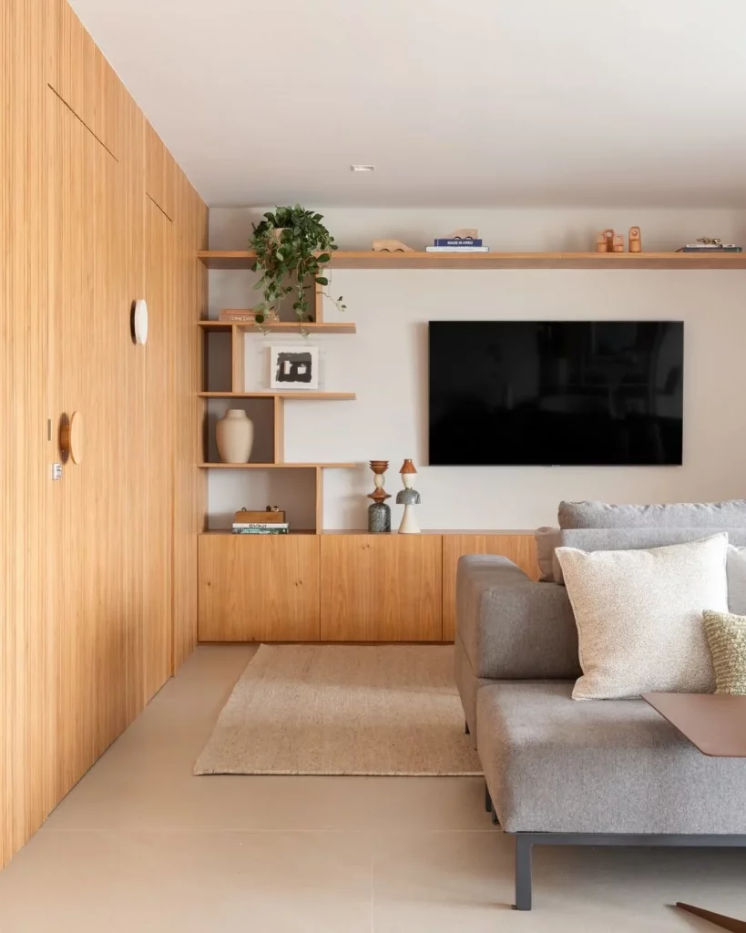 Prateleira de madeira com plantas, televisão na parede branca e sofá cinza.