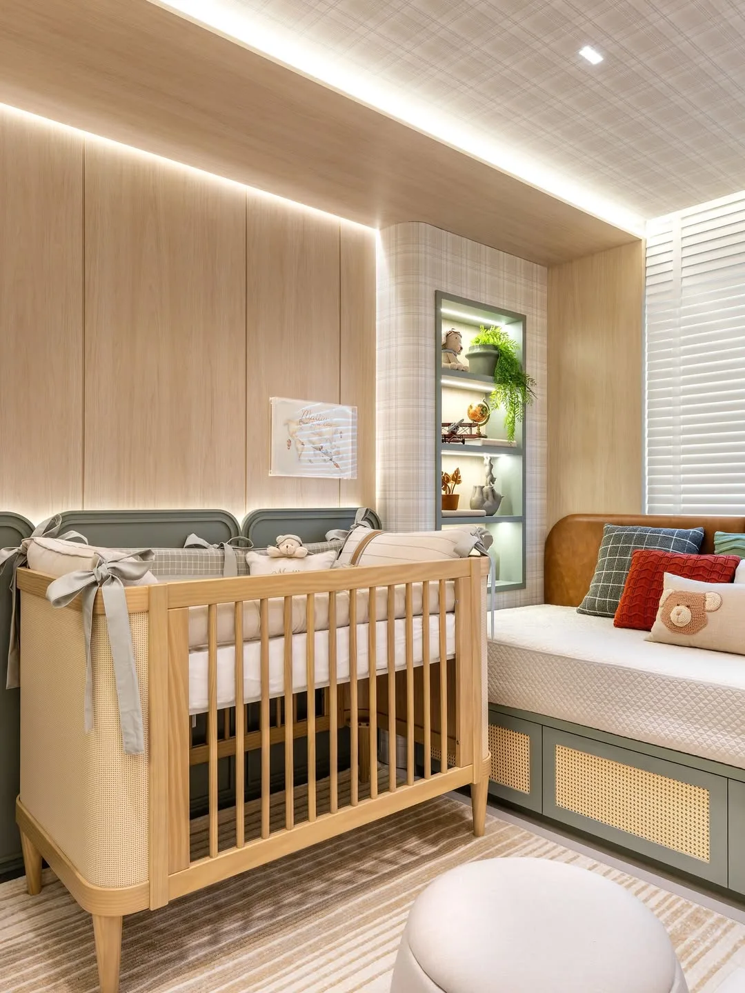 Quarto de bebê com berço de madeira clara, iluminação, almofada de leão no sofá.