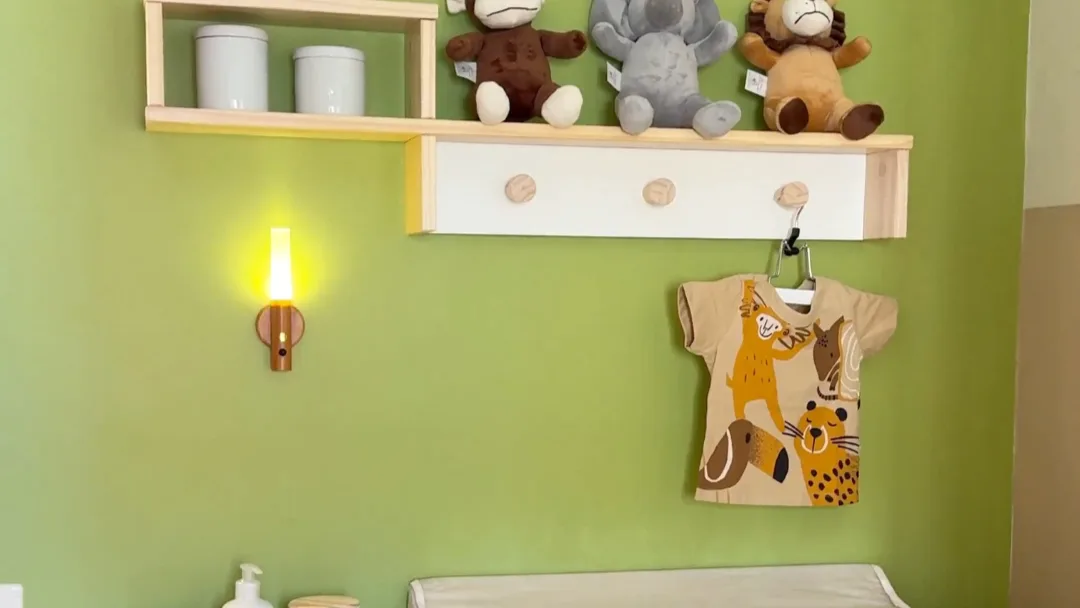 Parede verde no quarto infantil, animais de pelúcia, roupa pendurada e arandela ligada com luz quente.