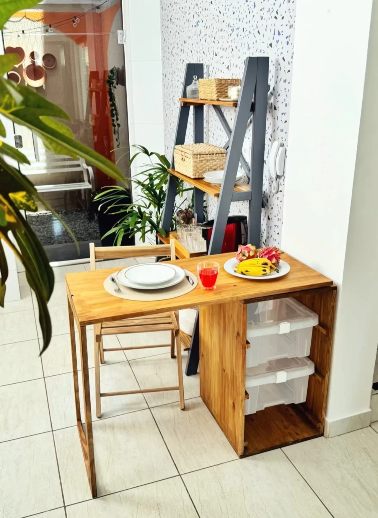 Cantinho do café com mesa de madeira, planta, artigos de mesa posta e estante no fundo.