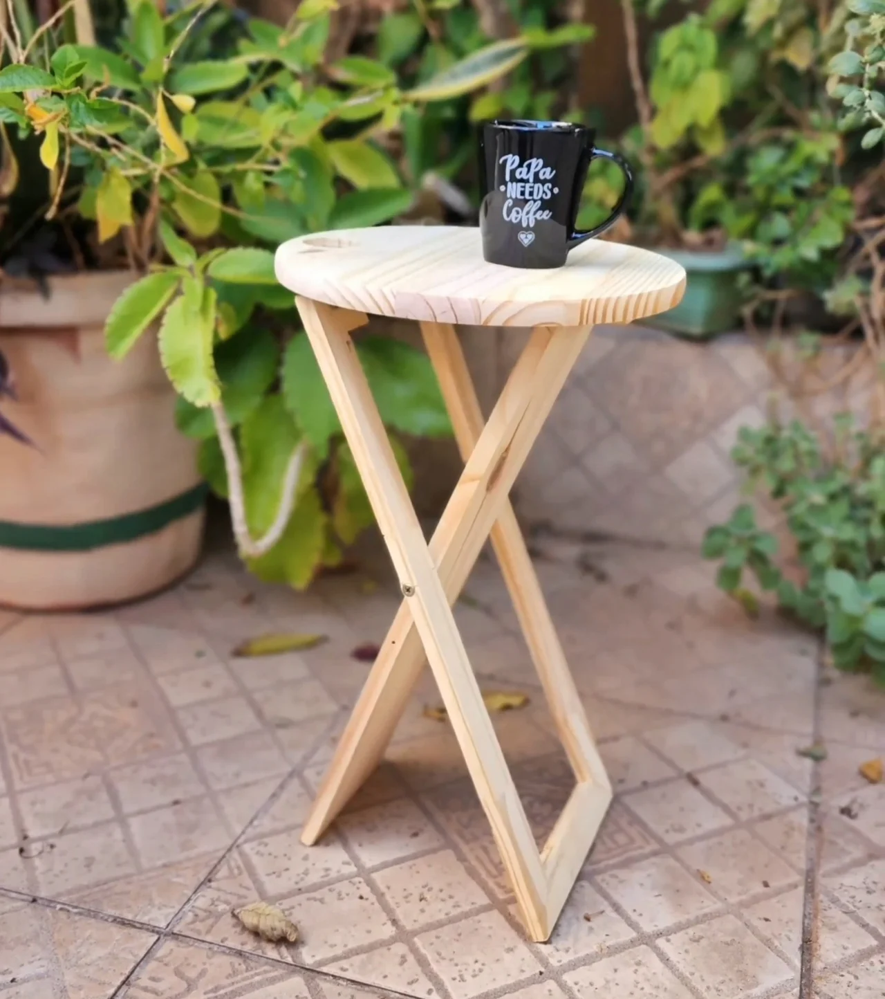 Mesa dobrável de madeira clara e caneca preta em cima, plantas no fundo.